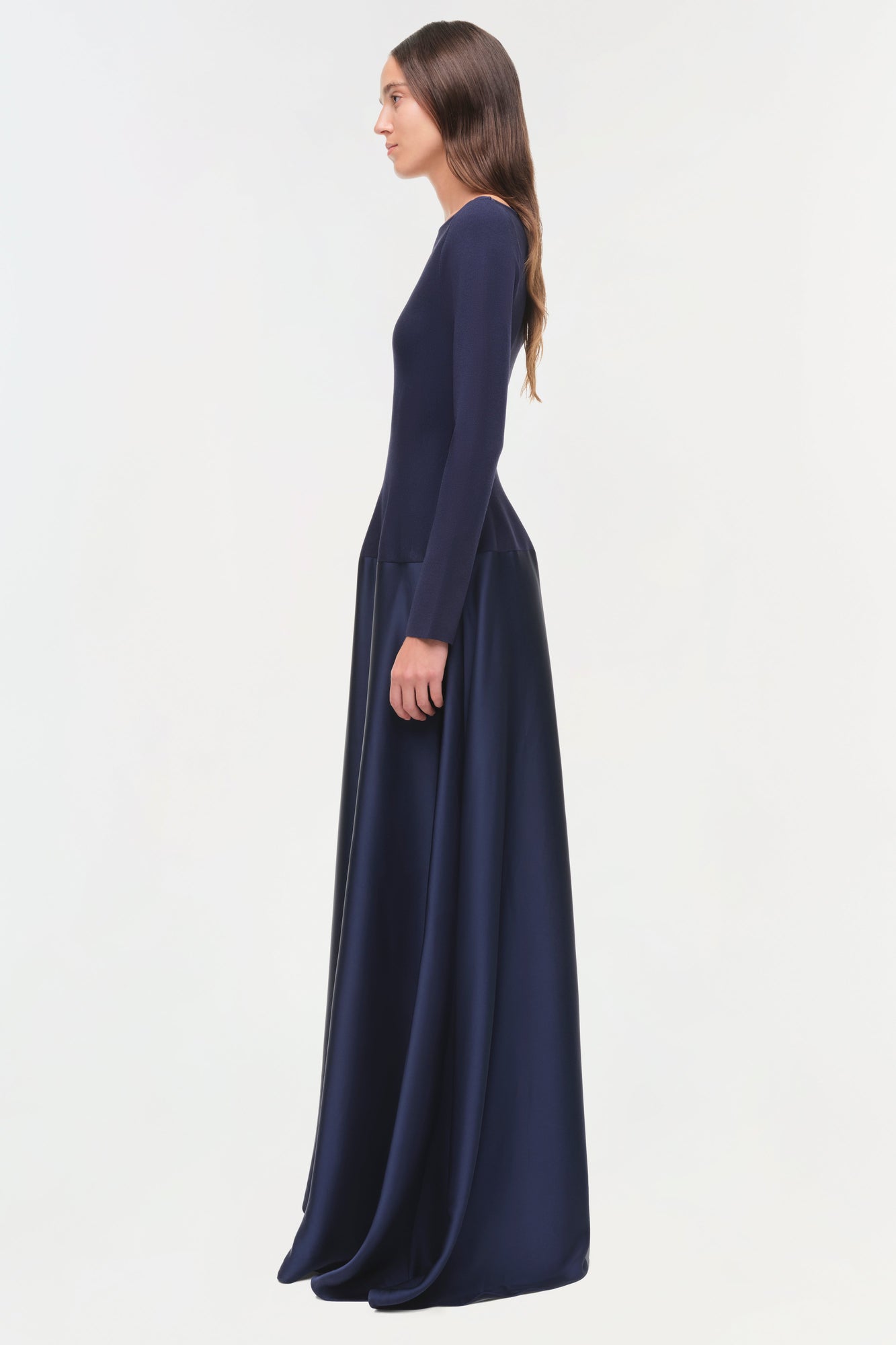 Woman wearing a navy satin knit gown with long sleeves and flowing skirt, shown in profile view.