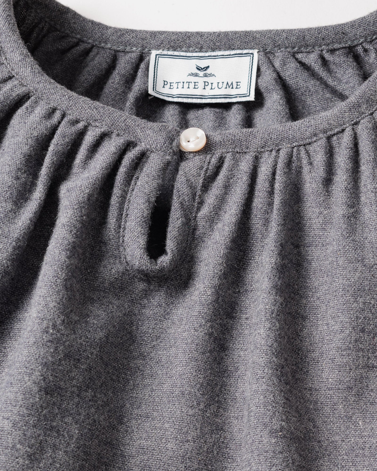 Close-up of a grey nightgown showcasing a round neckline and pearl-like button. Inside, the label reads Petite Plume with a bird graphic. Made from brushed cotton flannel, its texture highlights the softness and chemical-free comfort of the Girls Flannel Delphine Nightgown in Grey by Petite Plume.