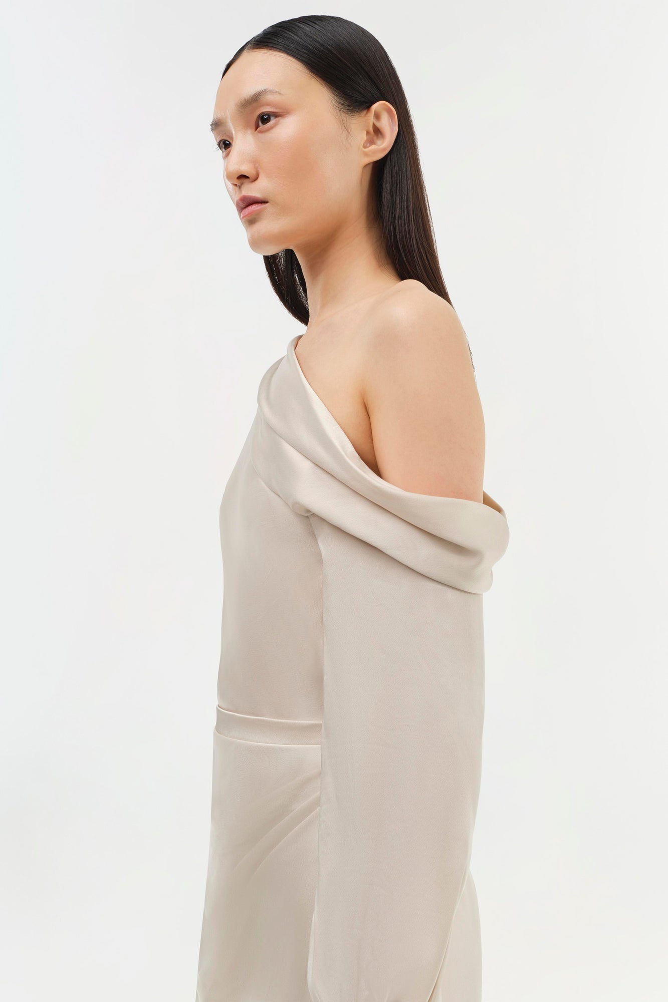 Cream-colored off-shoulder dress with draped neckline and long sleeves, shown on model in profile view.