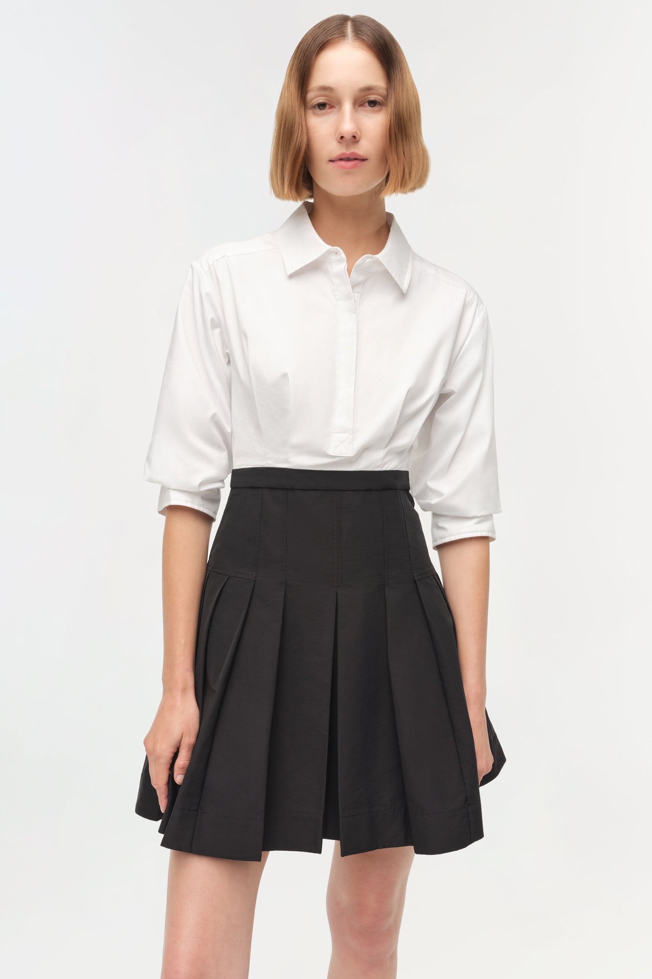 Woman wearing white button-up shirt with three-quarter sleeves over black pleated mini skirt.