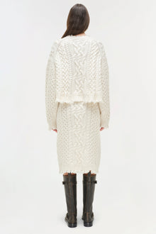 Cream cable-knit midi skirt with distressed hem, worn with matching cropped cardigan and black boots.