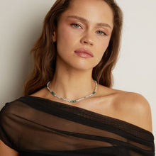 Model. Woman wearing a black off-shoulder top and green and silver necklace against a neutral background