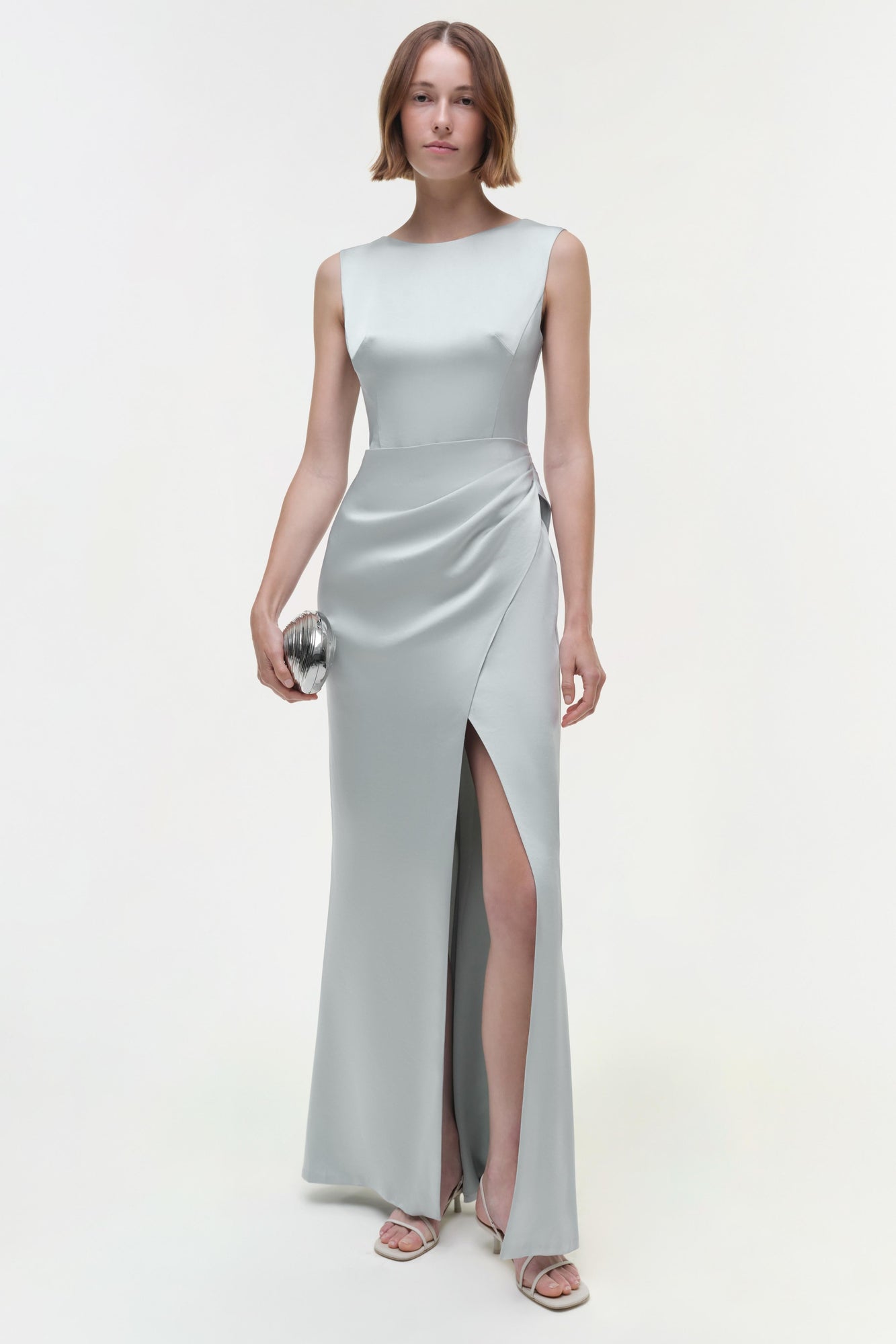 Light gray sleeveless gown with draped waist and high slit, shown on model wearing silver clutch.