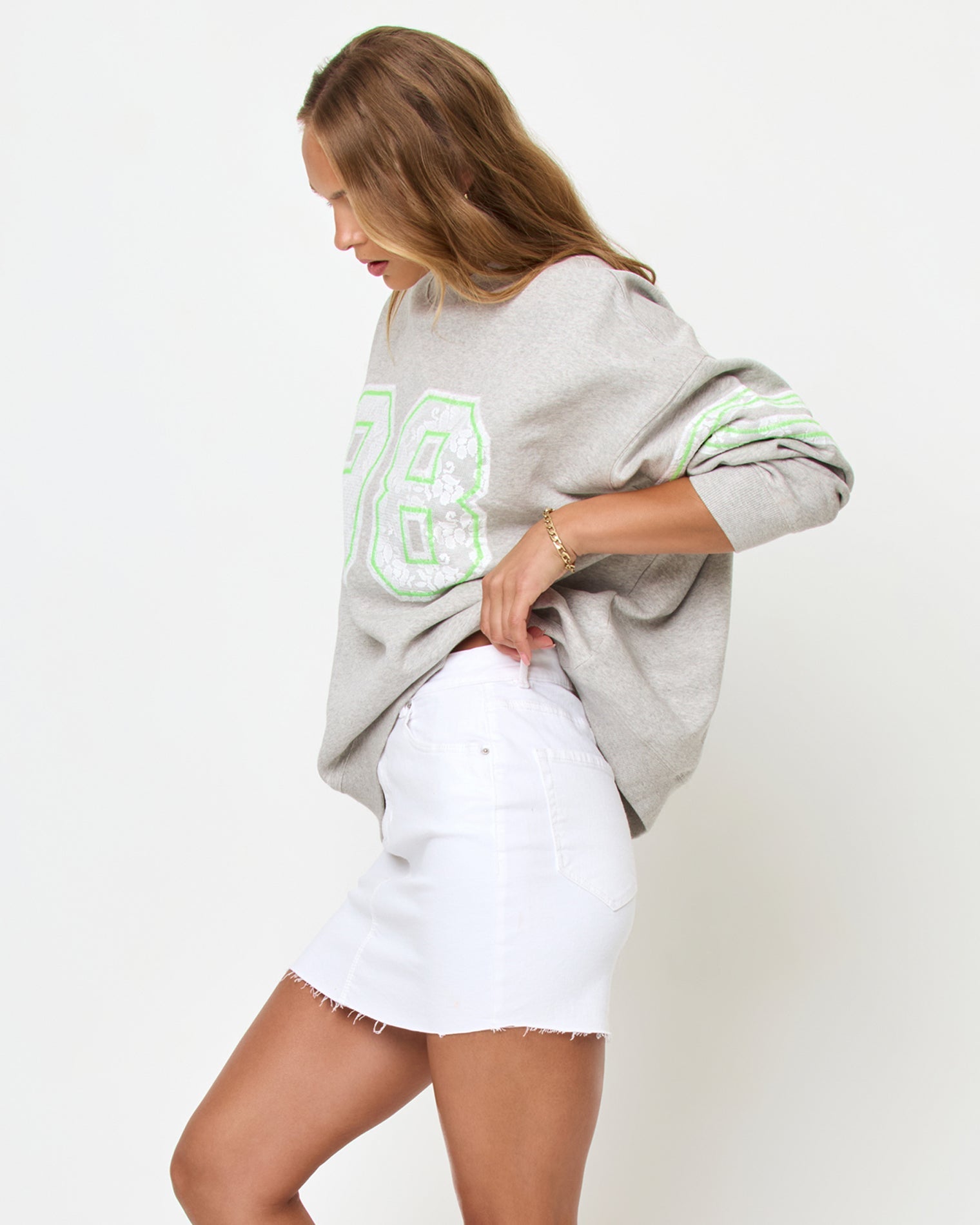 Angled side studio view of the Legend Sweatshirt in Limeade, a heather gray oversized crewneck sweatshirt with a neon green graphic, styled with a white denim skirt.