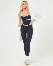 Front view of model wearing the Ace Jumpsuit in black-cream, a sleeveless jumpsuit with cream contrast piping, with white sneakers and an opened white button down shirt.