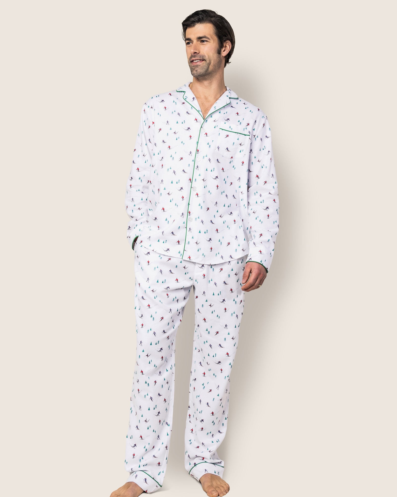A man models the Mens Flannel Pajama Set in Apres Ski by Petite Plume. Crafted from 100% cotton, it features a green-trimmed collar and cuffs. Against a plain background, he stands barefoot with one hand in his pocket, gazing slightly to the side.