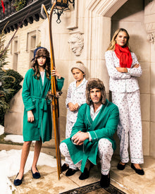 A family poses on a snowy porch in luxury sleepwear, wearing the Mens Flannel Pajama Set in Apres Ski by Petite Plume. One holds a ski, another sports a red scarf. A stone building and evergreen wreaths complete the cozy scene.