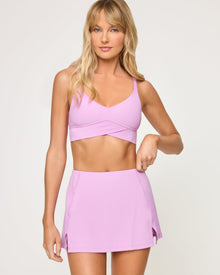 Close-up front view from the waist up of the LSPACE Unmatched Skort in mauve on a model against a white background, highlighting the cream crossover band detail and high waistband paired with the matching mauve crossover bra.