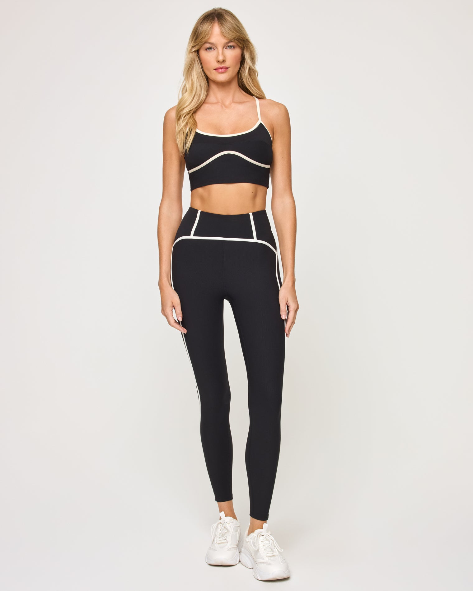 Front view of model wearing the Revel Legging in black-cream with cream piping trim and matching black-cream Revel Top, white sneakers.