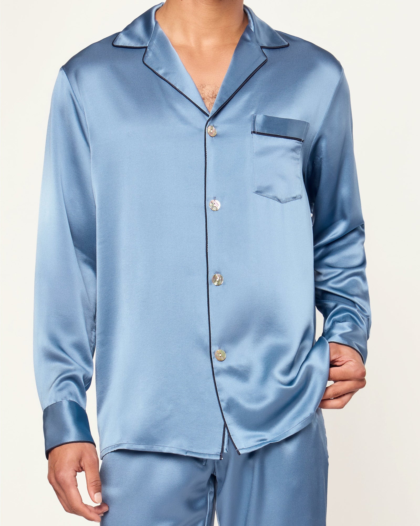The Men's Silk Pajama Set in Mystique Blue features a button-up long-sleeve top with a chest pocket and matching elastic-waist pants, both trimmed with dark piping.
