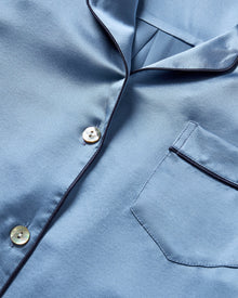 The Men's Silk Pajama Set in Mystique Blue features a button-up long-sleeve top with a chest pocket and matching elastic-waist pants, both trimmed with dark piping.