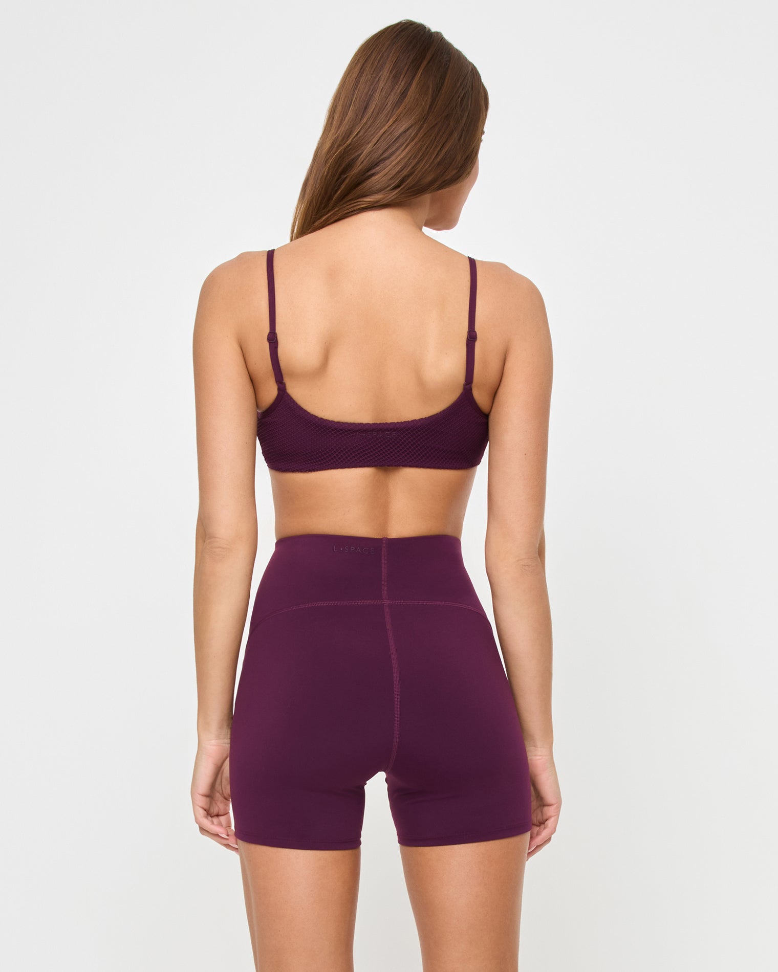 Back view of the LSPACE En Pointe Short in plum on a model against a white background, showing the back of the high-waist active shorts, paired with the matching plum Stride Bra.
