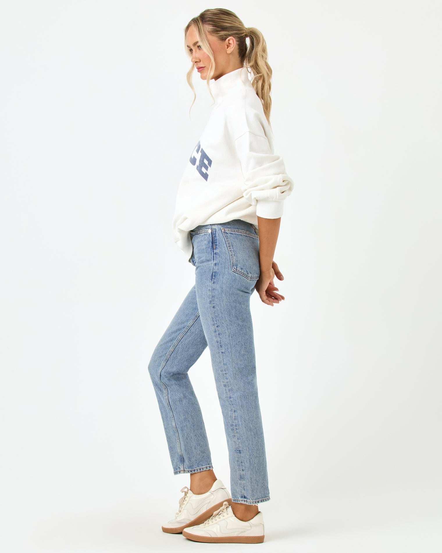 Side profile full-length studio view of the Crush Sweatshirt in cream, a cream quarter-zip pullover sweatshirt with "LSPACE" collegiate arch lettering, styled with blue jeans and white sneakers.