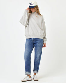 Front full-length studio view of the Crush Sweatshirt in Heather Gray, a heather gray oversized quarter-zip sweatshirt, styled with cuffed blue jeans, Adidas sneakers, and a navy and cream baseball cap.