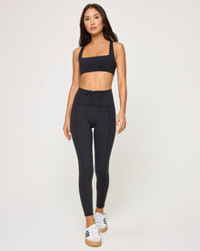 Front view of the LSPACE Uppercut Bra in black on a model against a white background, showing the square-neck sports bra with thick straps, paired with matching black high-rise active pants.