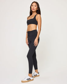 Side view of the LSPACE Uppercut Bra in black on a model against a white background, paired with matching black high-rise active pants, showing the profile of the square-neck bra design.