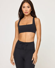 Close-up front view from the waist up of the LSPACE Uppercut Bra in black on a model against a white background, showing the square neckline and thick shoulder straps, paired with matching black high-rise active pants.