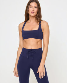 Close-up front view from the waist up of the LSPACE Uppercut Bra in midnight blue on a model against a white background, showing the square neckline and thick straps, paired with matching midnight blue high-rise active pants with lace-up waistband detail.
