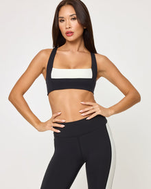 Lifestyle editorial shot of a model wearing the LSPACE Uppercut Bra in black-cream paired with matching black-cream high-rise active pants, in an outdoor setting.