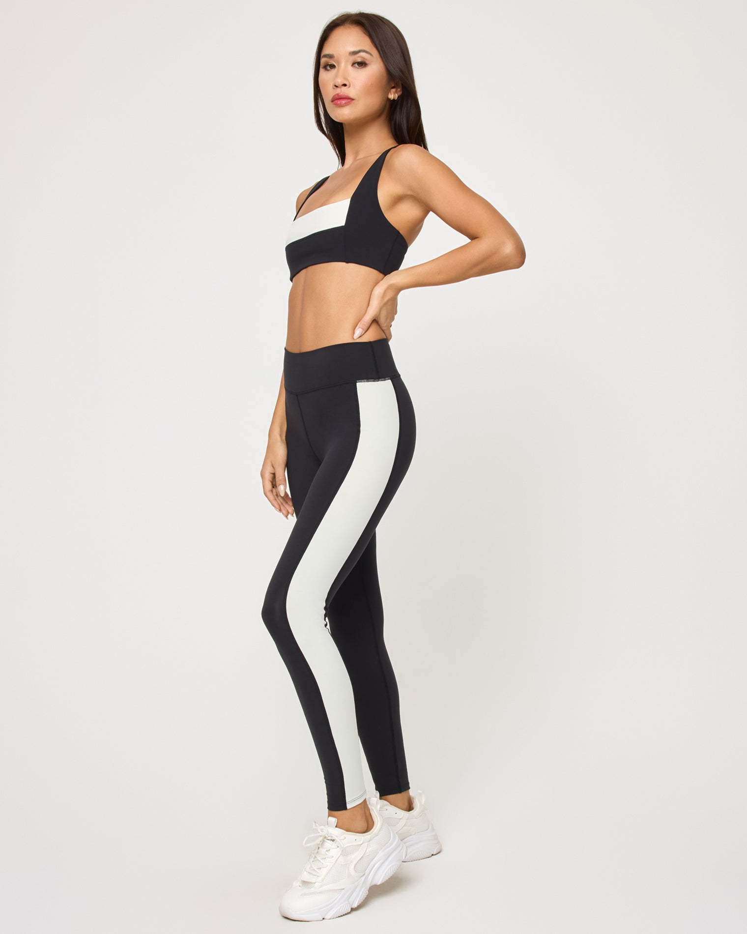 Side view of the LSPACE Uppercut Bra in black-cream on a model against a white background, paired with black-cream high-rise active pants, showing the profile of the matching outfit.