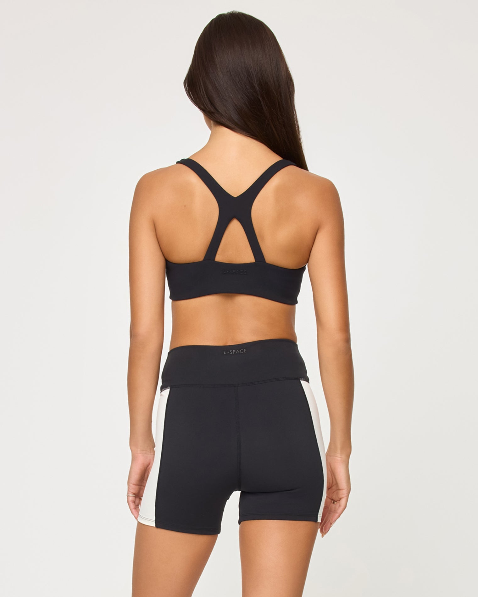 Back view of the LSPACE Uppercut Short in black-cream on a model against a white background, showing the back of the high-rise active shorts with cream trim detail, paired with the matching black-cream sports bra.