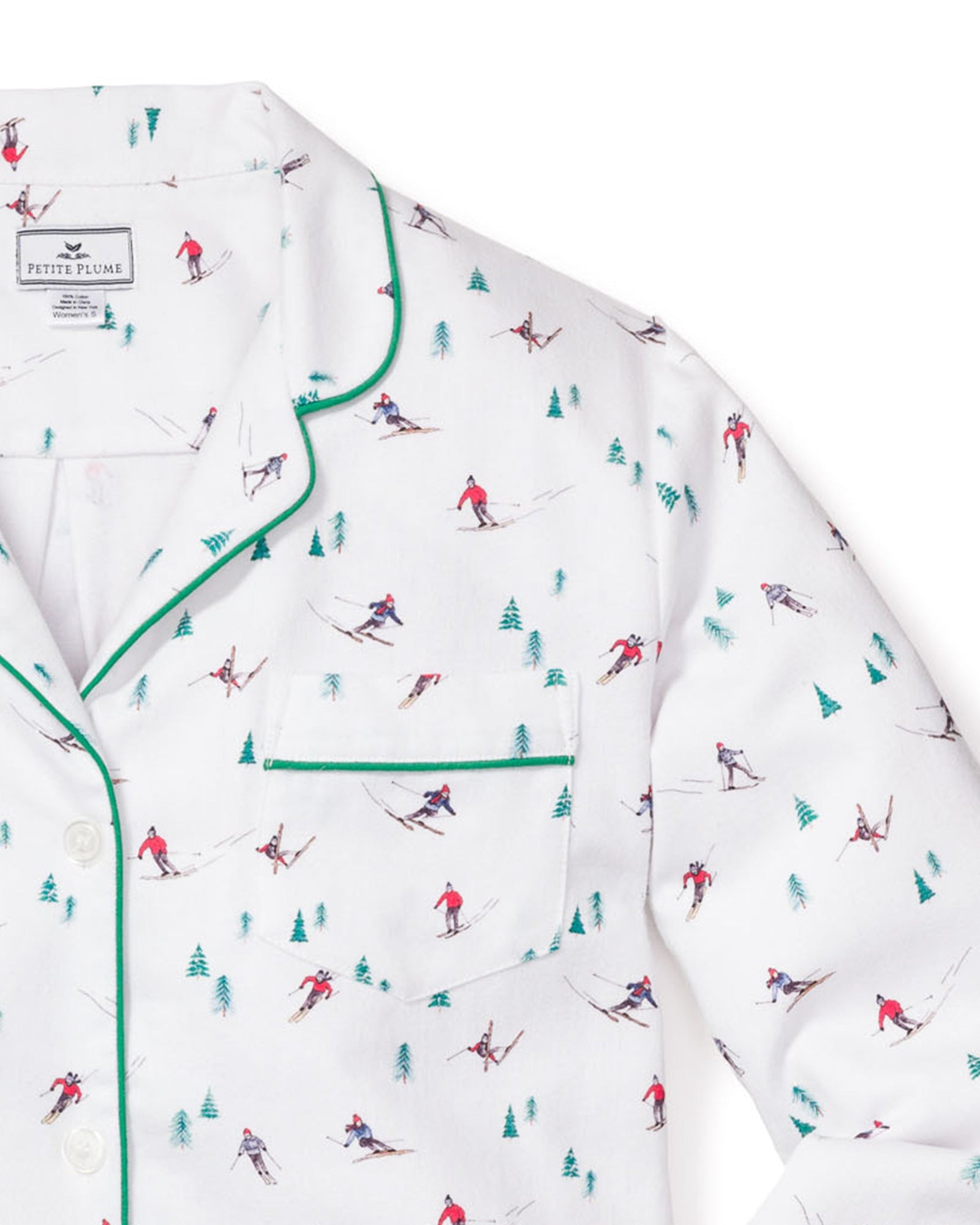 The Womens Flannel Pajama Set in Apres Ski by Petite Plume is a white brushed cotton flannel top featuring a vibrant skier and pine tree pattern, accented with green piping on the collar and pocket for stylish comfort.