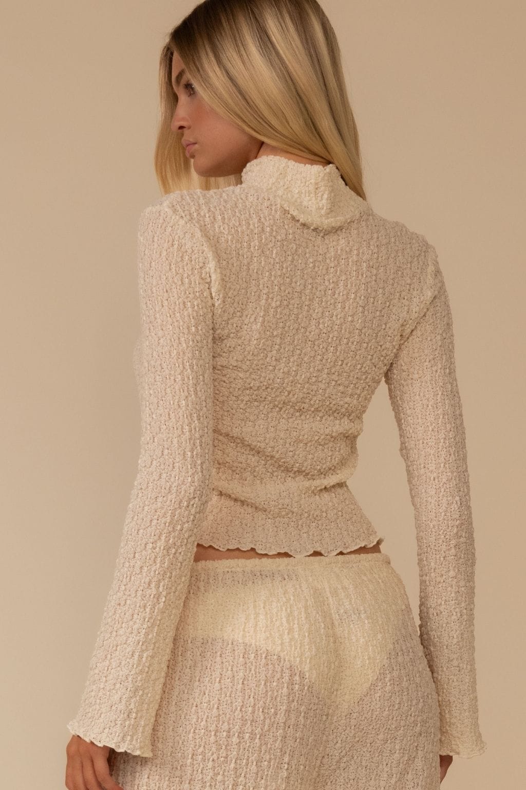 Cream textured long-sleeve top with cowl neckline and fitted waist, shown on model from back view.