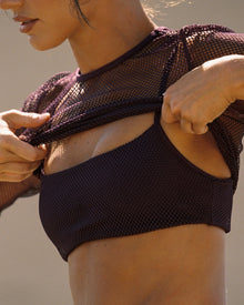 Close-up editorial detail shot of a model adjusting the LSPACE Stride Bra in plum under a plum mesh fishnet long-sleeve top, showing the upper body and bra styling.