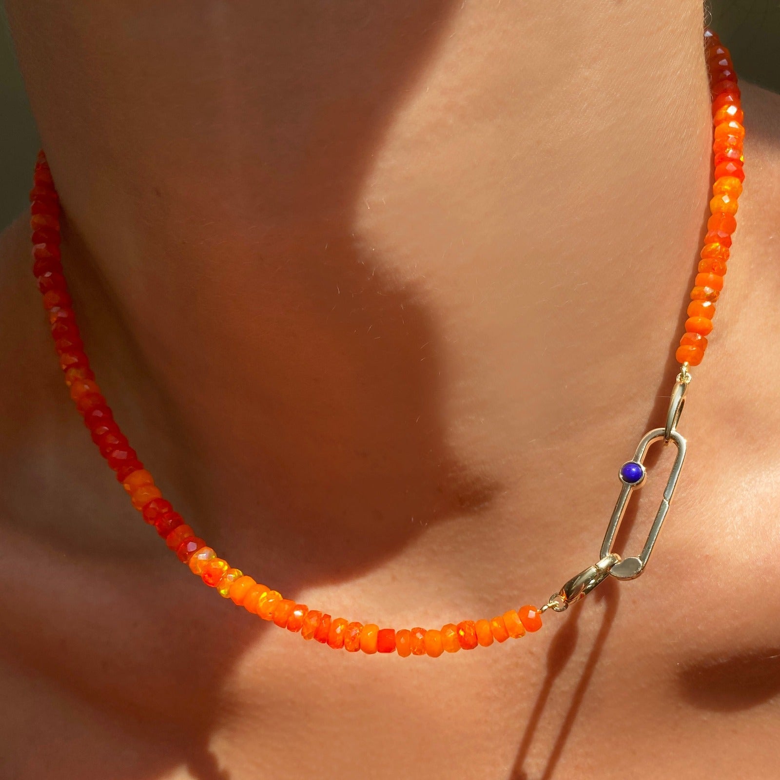 Shimmering beaded necklace made of faceted opals in shades of orange on a gold linking ovals clasp. Styled on a neck layered with a paperclip charm lock.