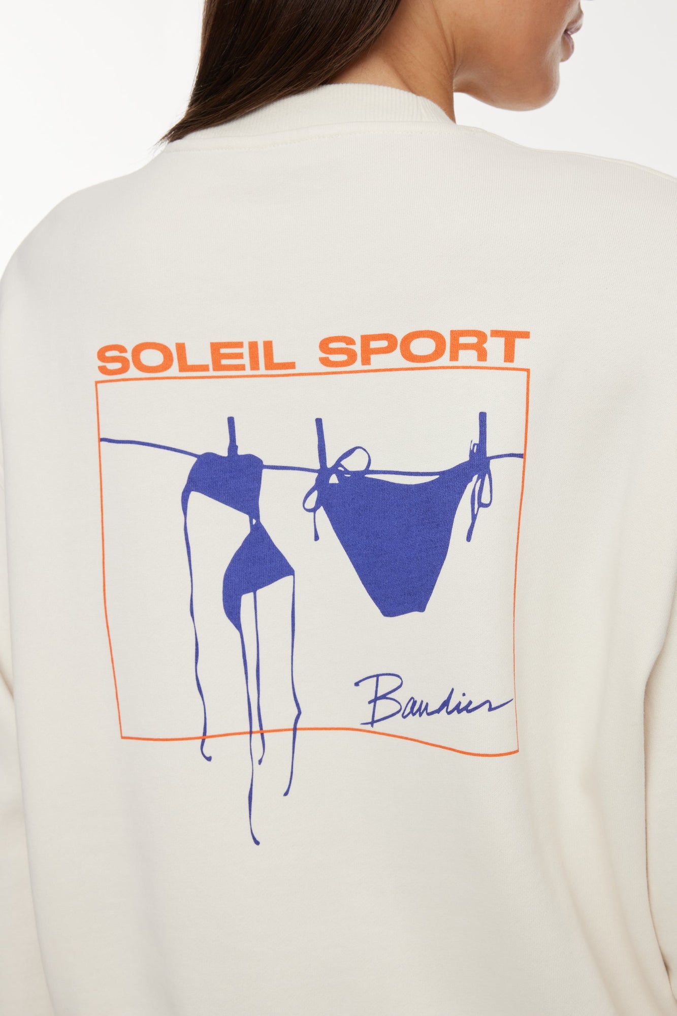 Model wears a white crewneck sweatshirt with logo on back that reads "Soleil Sport" and underneath is a square orange border containing a blue bikini hanging to dry on a clothesline. 