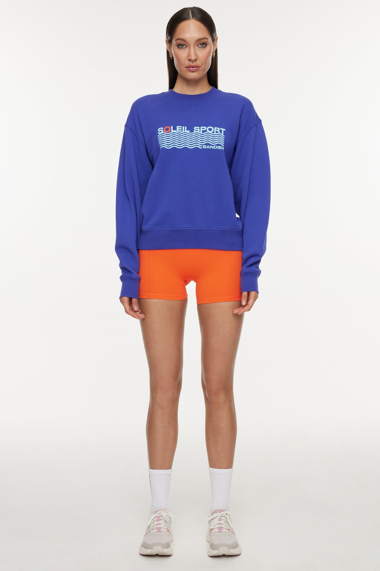 Model wears a royal blue crewneck sweatshirt with the lettering "Soleil Sport" across the chest with wavey lines below and "Bandier" in the bottom right corner. 