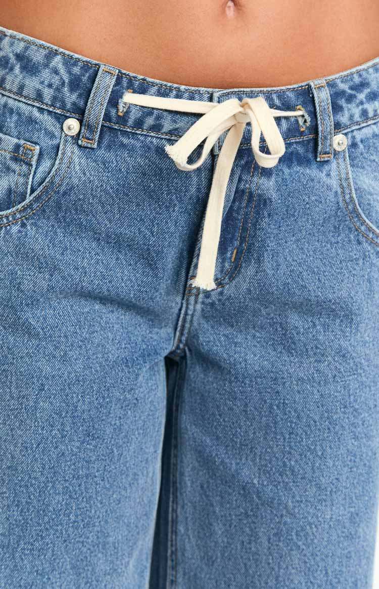 Close-up of the cream tie waist drawstring, tied in a bow, with distressed opening on mid wash denim jeans