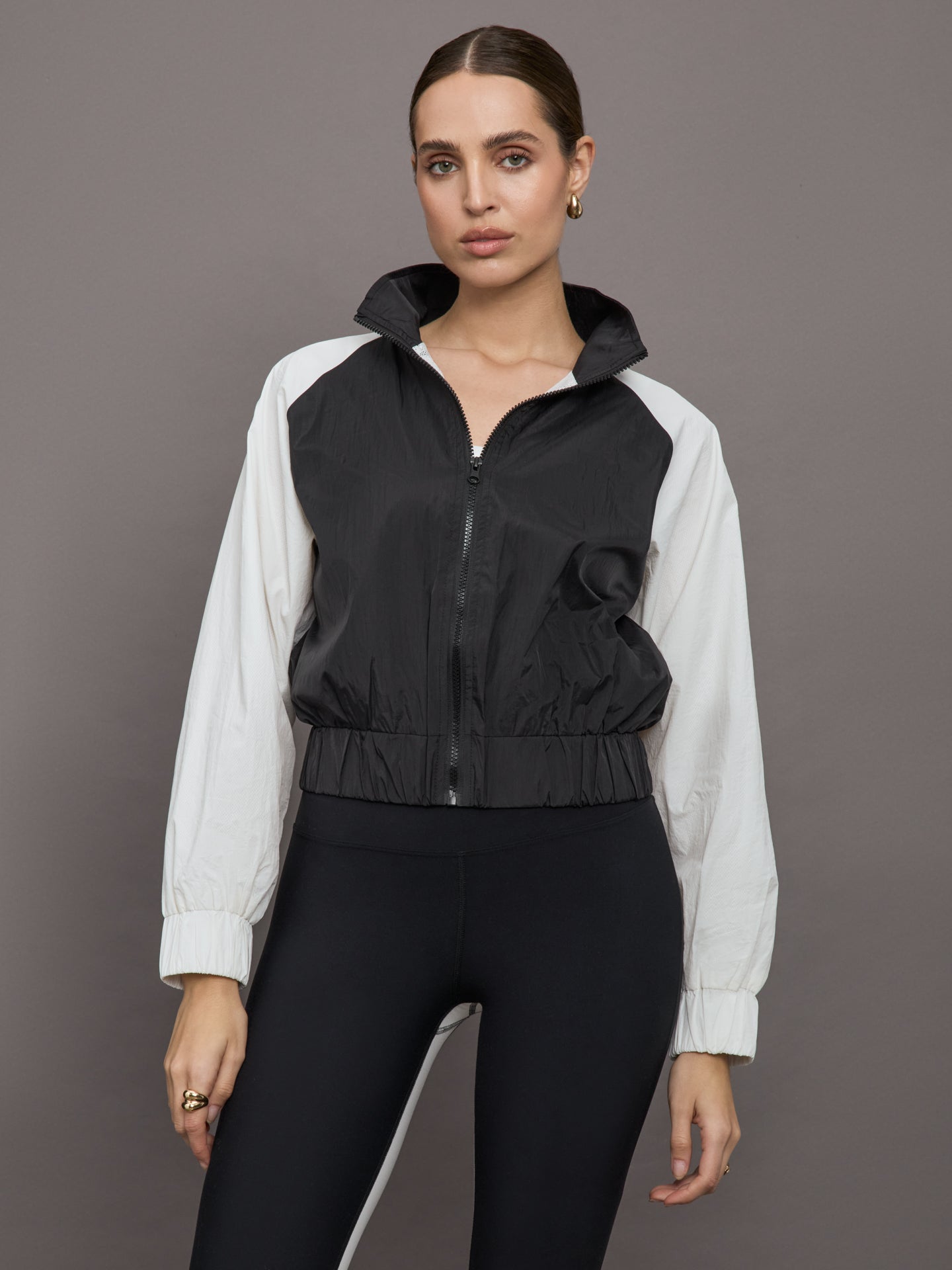 Model wears a cropped windbreaker with a popped collar, black bodice and white long sleeves with a cinched wristline.