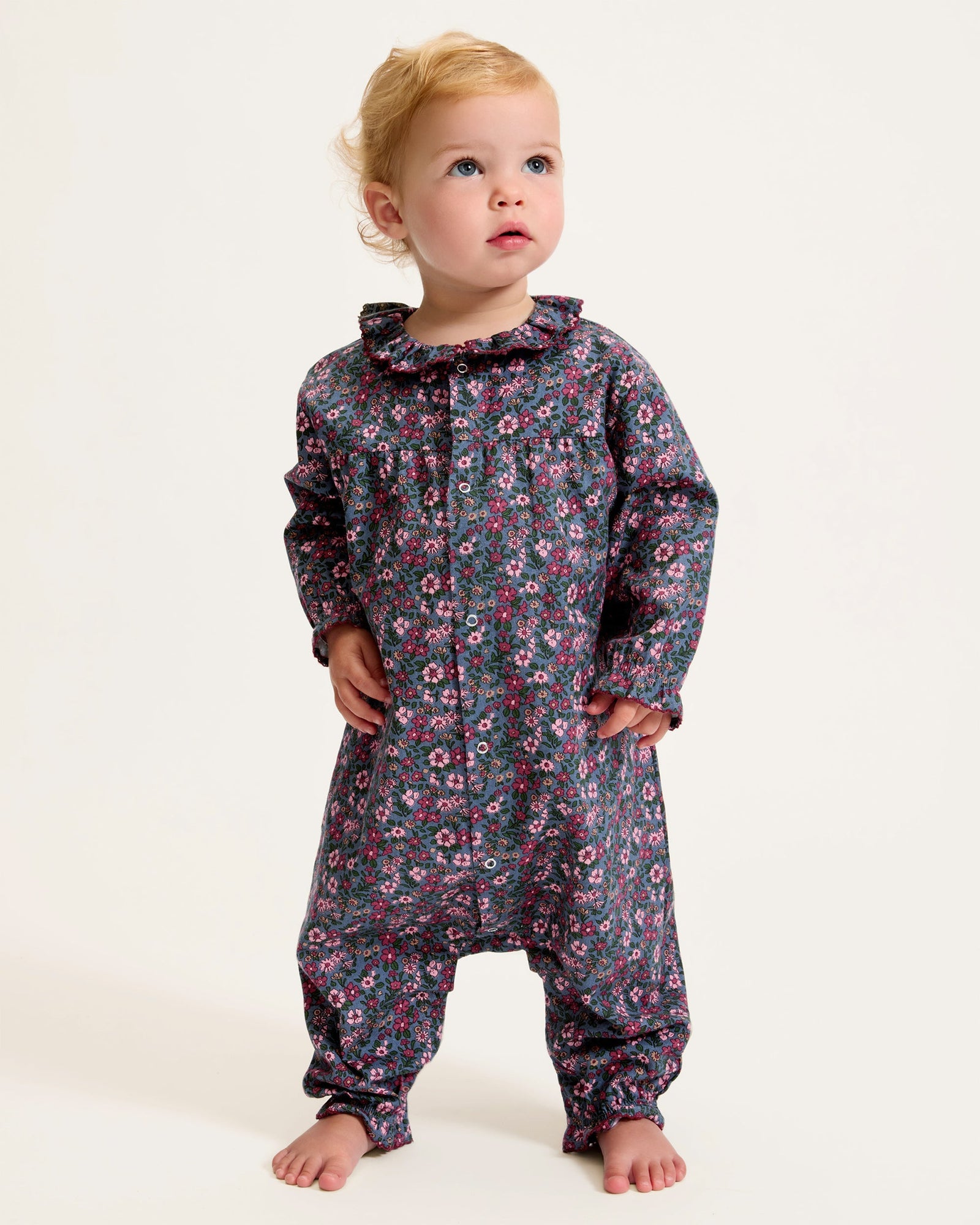 Baby's Twill Madeline Romper in Fleurs de Saison features long sleeves, a dark base with pink and purple floral print, front snap closures, a rounded collar, and gathered cuffs at wrists and ankles.