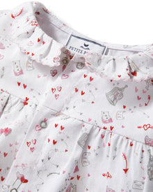 The Baby's Twill Madeline Romper in Love Song is a white long-sleeve romper with ruffled cuffs and collar, decorated with a pink pattern of hearts, glasses, and small animals. It features snap buttons down the front and legs.