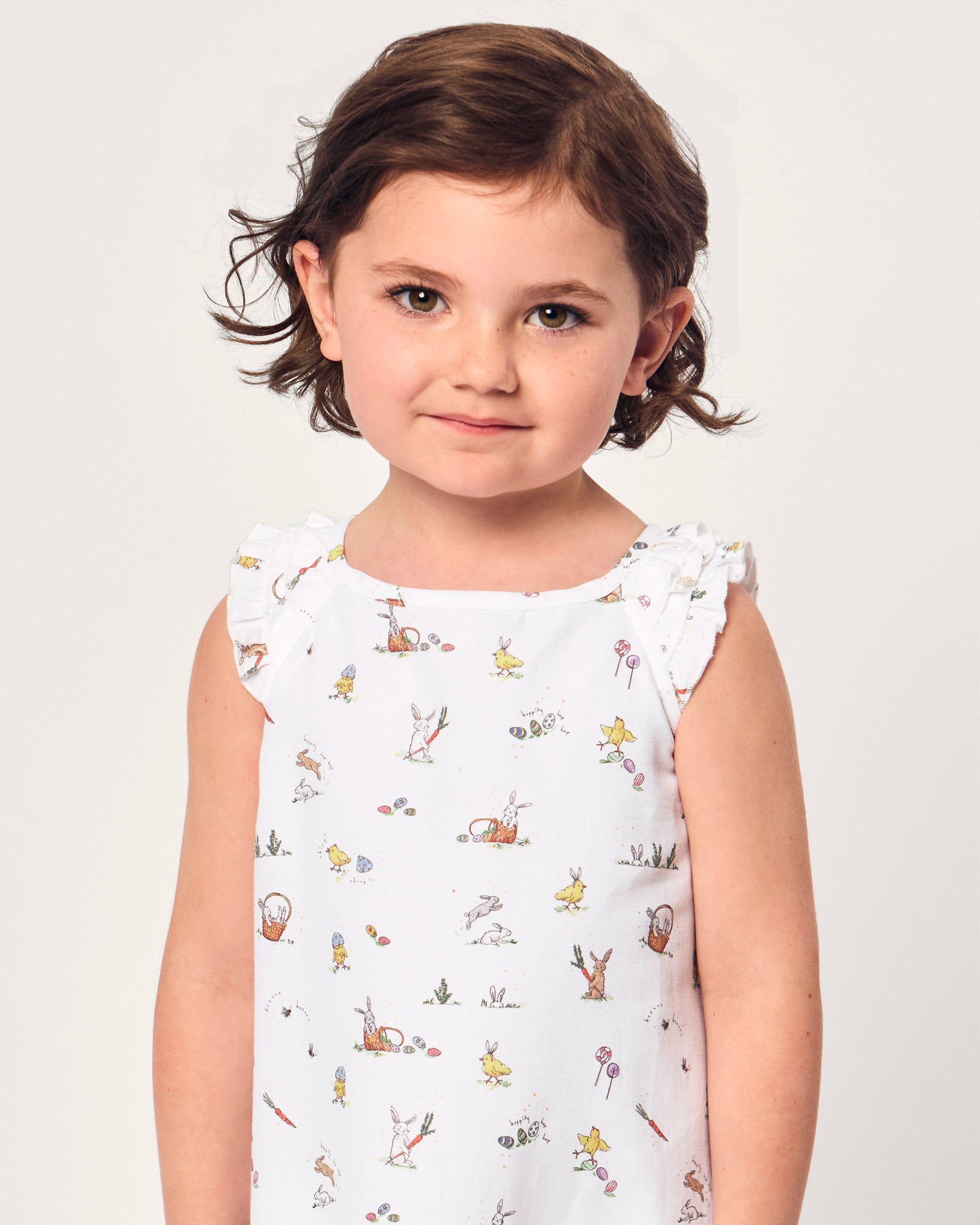 A young girl with short brown hair smiles in the Girl's Twill Amelie Nightgown in Easter Frolic, a white sleeveless dress featuring playful animal and nature prints, standing against a plain light background.