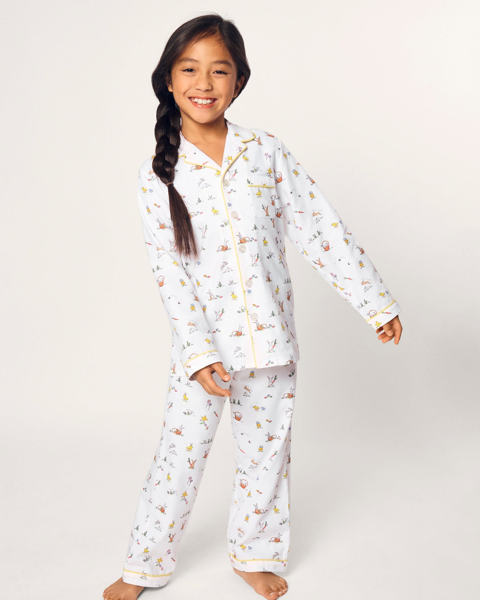 A young girl with long dark hair in a braid smiles barefoot, wearing the Kid's Twill Pajama Set in Easter Frolic, against a plain light background.
