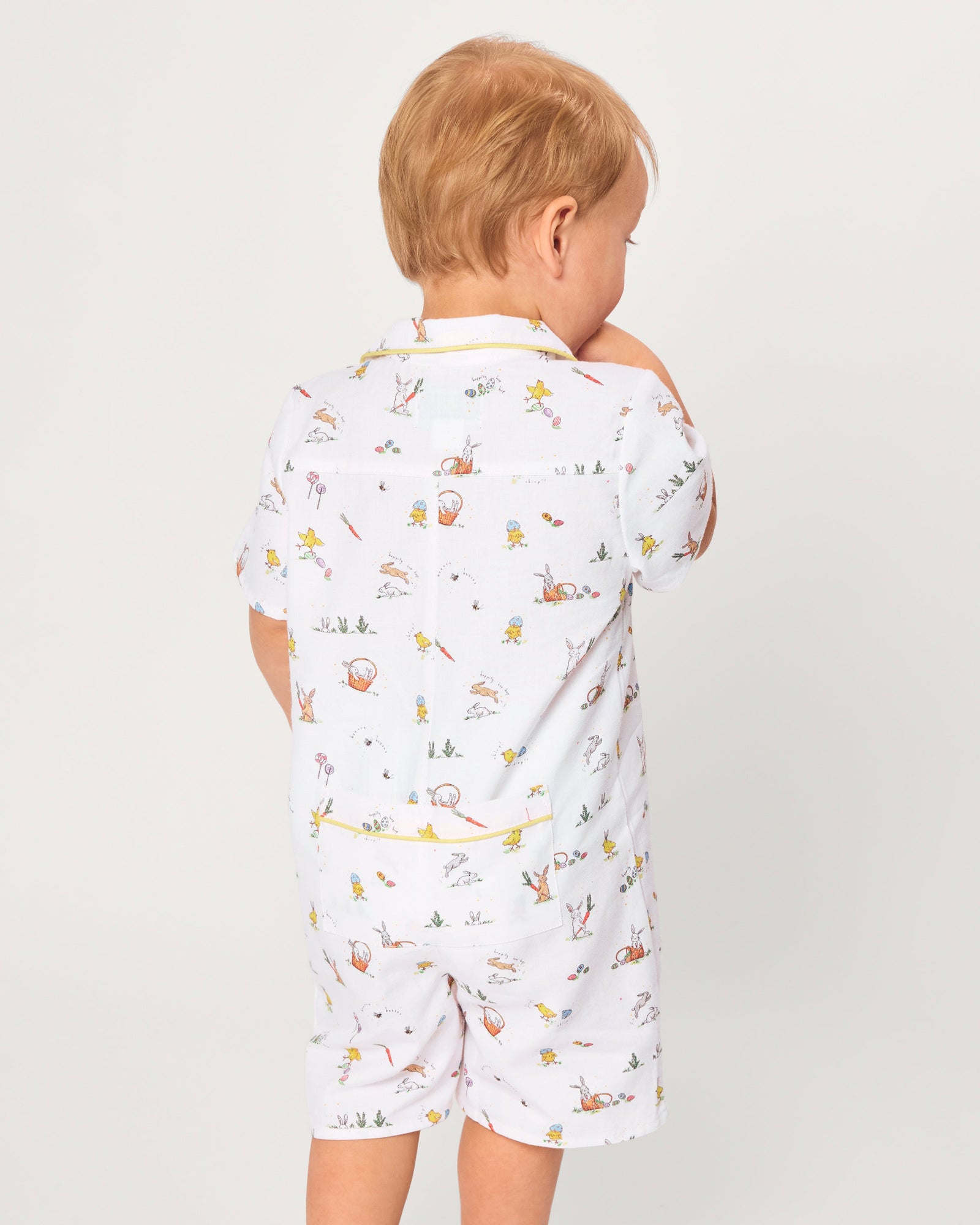 A young child with blonde hair wears the Baby's Twill Summer Romper in Easter Frolic—white with colorful animal prints and yellow trim—standing against a plain light background, holding two wooden blocks and looking to the side.