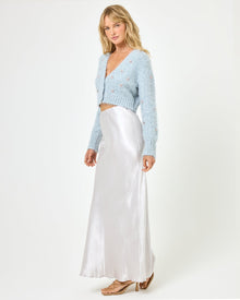 Three-quarter side view of a model wearing the LSPACE Sweet As Honey Skirt in silver, showing the high waist and fluid satin drape, styled with a pale blue embellished cardigan on a white background.