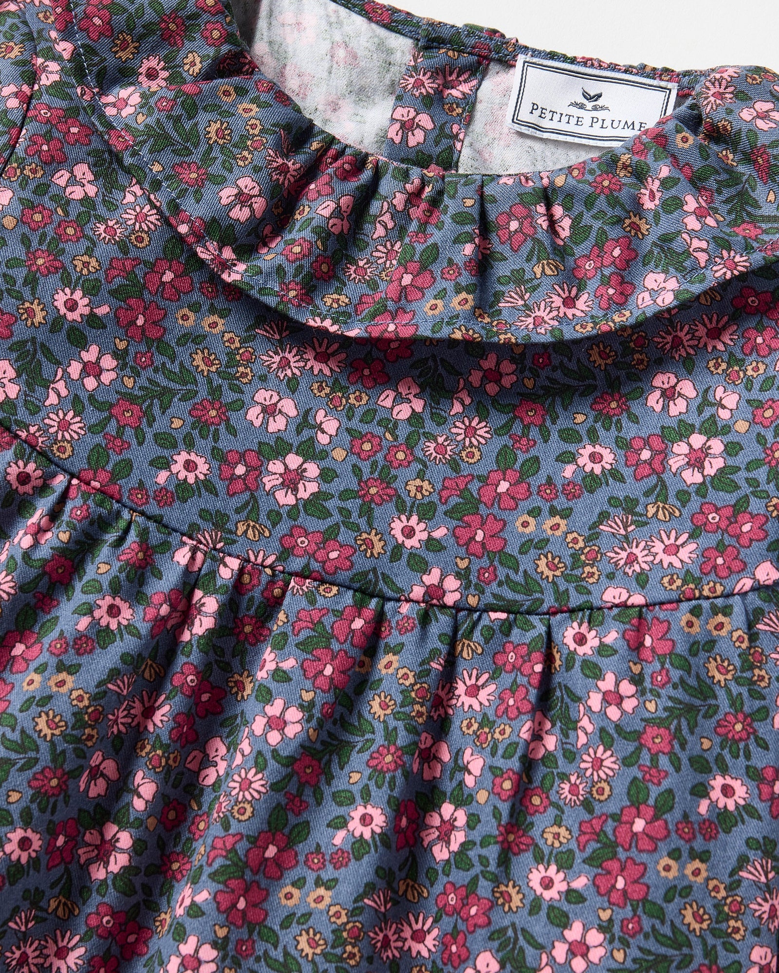 The Girl's Twill Scarlett Nightgown in Fleurs de Saison is a long-sleeved, knee-length dress with a ruffled collar and cuffs, featuring pink and white flowers with green leaves on a dark background.