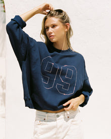 Lifestyle editorial shot of a model wearing the LSPACE Legend Sweatshirt in midnight blue, an oversized cropped sweatshirt with a rhinestone number graphic, paired with white denim jeans against an outdoor white wall.