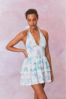 White halter dress with a blue floral pattern, halter ties in bow at neck and below, is a wide smocked waistband and the skirt has two ruffled tiers.