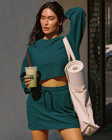 Model outdoors on a city street wearing the Half Court Pullover in deep teal, a ribbed long-sleeve cropped crewneck, with matching deep teal Half Court Skirt, holding a green drink and carrying a yoga mat bag, Adidas sneakers.