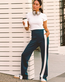 Model outdoors on an urban street wearing the Back To Back Pant in deep teal-cream, a wide-leg track pant with cream contrast side stripe, with matching deep teal-cream Back To Back Jacket and white sneakers.