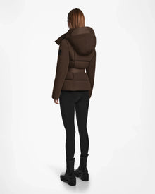 A person with straight, shoulder-length hair stands facing away, wearing the Rudsak ADELINE - 8124544 Mocha quilted short down puffer, black leggings, and black ankle boots against a plain white background.