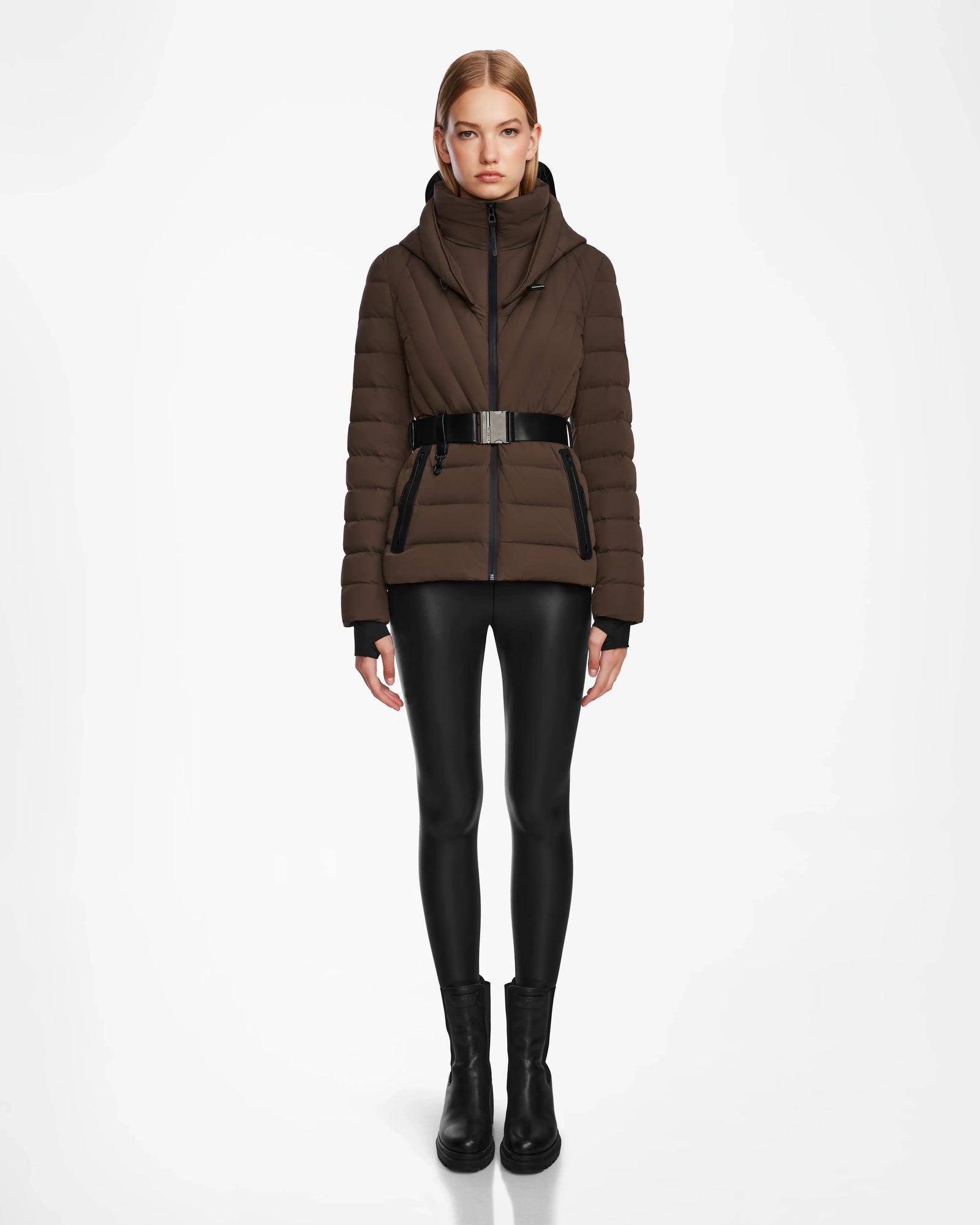 A woman stands facing forward in the Rudsak ALISA - L124506 Espresso womens quilted down puffer jacket with a vegan leather belt, paired with black leggings and boots, against a plain white background.