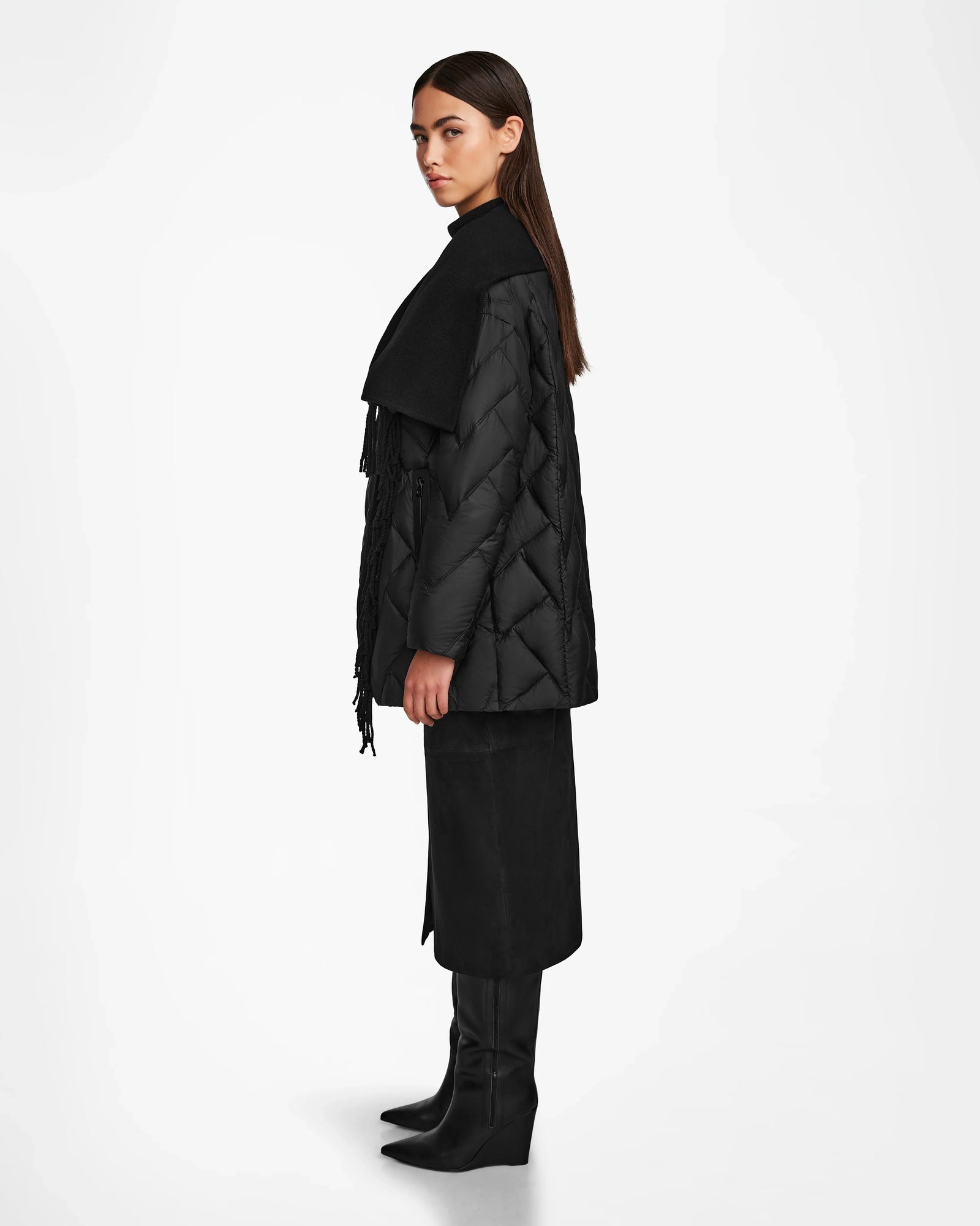 A woman stands in profile against a plain white background, wearing the Rudsak ANNE MARIE - 8124713 Black mid-length jacket, paired with a black scarf, black midi skirt, and knee-high black boots.