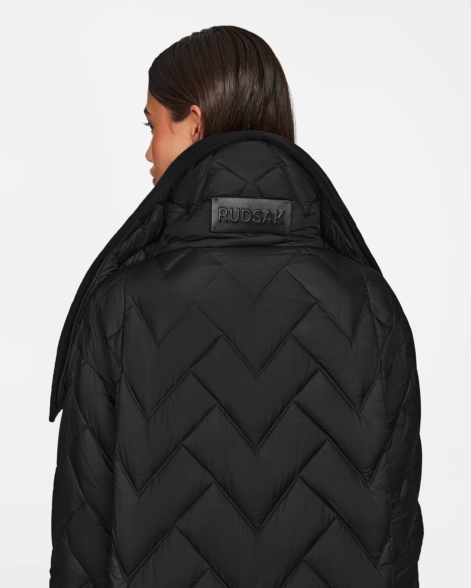 A woman with straight brown hair wears the Rudsak ANNE MARIE - 8124713 Black mid-length puffer, featuring a chevron pattern, high collar, and embossed leather RUDSAK logo patch. She is shown from the back.