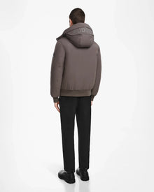 A person stands facing away, wearing the Rudsak ASH - 6124599 Grey water-resistant bomber with RUDSAK on the hood, black pants, and black shoes against a plain white background.