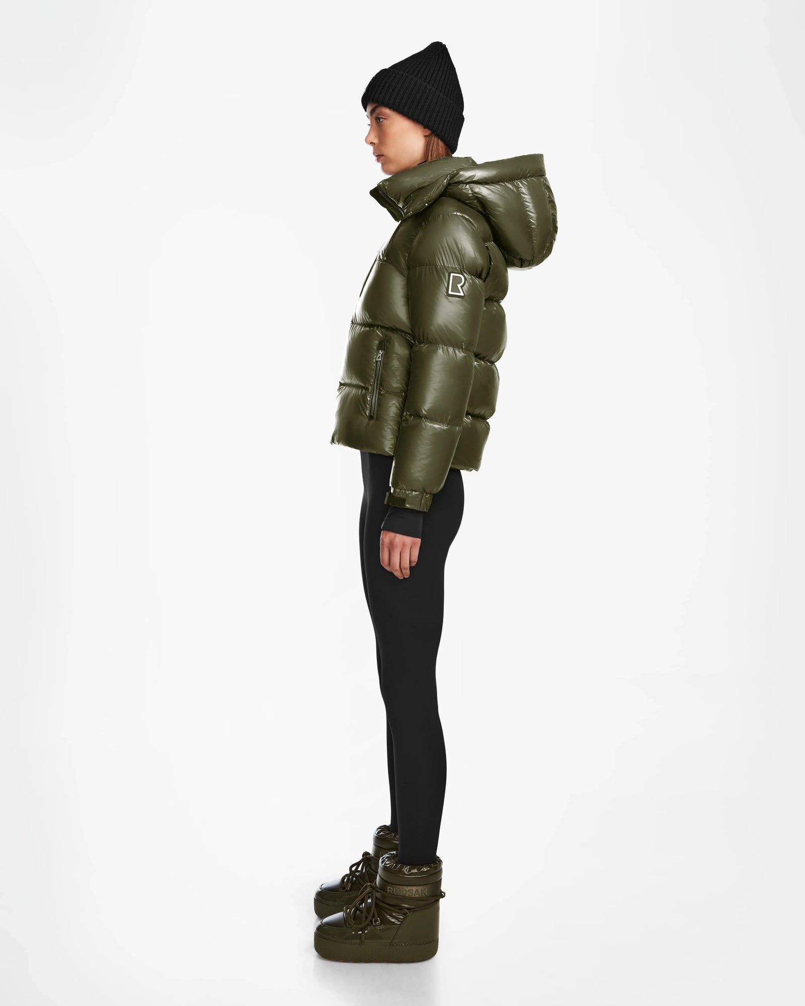 A woman in profile wears the Rudsak ASHLYN S - 8124565 Loden Green water-resistant down jacket with hood, black beanie and leggings, plus olive green winter boots, standing against a plain white background.